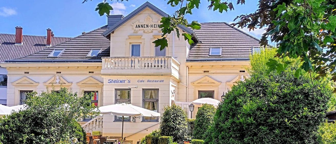 Das Äußere eines großen Gebäudes namens 'ANNEN-HEIM' zeigt einen weitläufigen Rasen mit einer Holzbank, Sträuchern und einer Gartenmauer. Vor dem Gebäude sind Sonnenschirme aufgestellt.