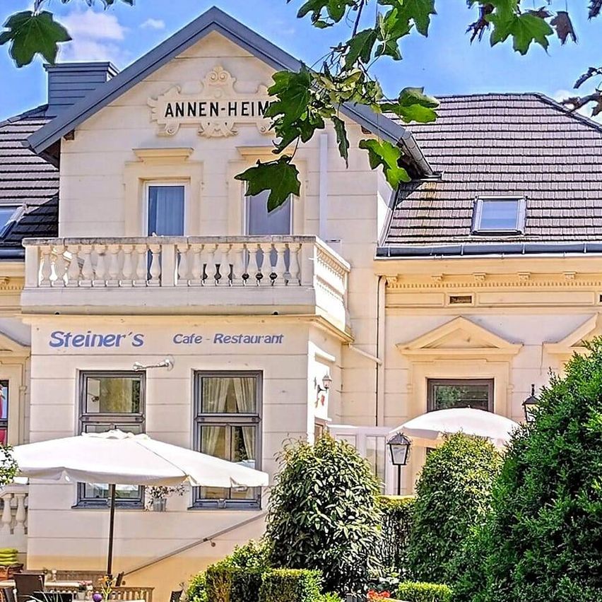 Das Äußere eines großen Gebäudes namens 'ANNEN-HEIM' zeigt einen weitläufigen Rasen mit einer Holzbank, Sträuchern und einer Gartenmauer. Vor dem Gebäude sind Sonnenschirme aufgestellt.