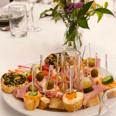 Ein weißer Tisch mit verschiedenen Speisen auf einem Teller, einschließlich Oliven, Käse und Gemüse. Eine Vase mit Blumen ist ebenfalls vorhanden.