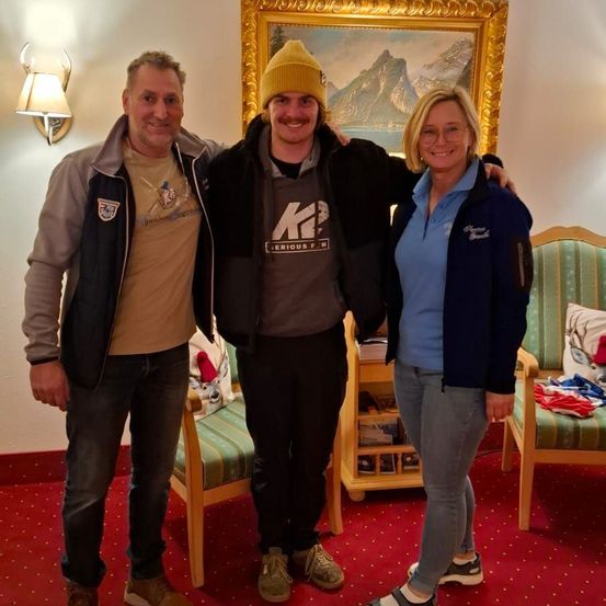 Drei Personen lächeln für ein Foto in einem Raum mit rotem Teppich, einer an der Wand montierten Lampe und einem gerahmten Gemälde. Hinter ihnen steht ein Holzstuhl mit einem Kissen.