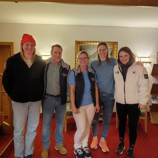 Fünf Menschen stehen zusammen in einem Raum. Sie alle lächeln. Eine Frau mit Brille und blauer Jacke steht in der Mitte. Ein Mann in einem karierten Hemd steht links. Eine Frau mit einer roten Mütze steht rechts.