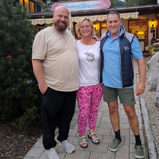 Drei Personen posieren für ein Foto vor einem Gebäude mit einem Schild, auf dem 'Alpenhotel Bergzauber' steht. Ein Mann und eine Frau, lässig gekleidet, stehen auf einem Weg mit einem Baum und Felsen im Hintergrund.