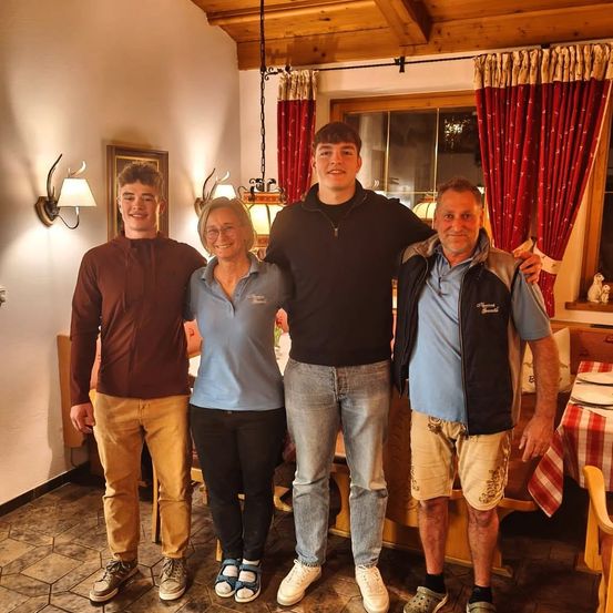 Vier Menschen stehen in einem Esszimmer zusammen. Zwei Männer, einer in einem braunen Jackett und der andere in einem schwarzen Jackett, flankieren eine Frau in einem blauen Shirt. Dahinter ist ein Esstisch mit Tellern gedeckt.