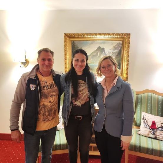 Drei Menschen stehen in einem Raum mit einem Bilderrahmen und lächeln. Einer trägt eine Jacke und Hose, ein anderer eine Jeansjacke und Leggings und der dritte einen Blazer. Hinter ihnen steht ein Stuhl mit einer Kissen.