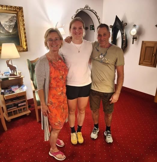 Drei Personen stehen auf einem roten Teppich. Eine Frau in einem orangefarbenen Kleid, eine Frau in einem weißen T-Shirt und ein Mann in einem grünen T-Shirt. Hinter ihnen steht ein grüner Stuhl und eine Lampe.