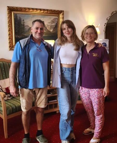 Drei Menschen stehen in einem Zimmer mit Teppich und lächeln. Ein Mann in einem blauen Shirt und Shorts, eine Frau in einem weißen Crop-Top und Jeans und eine weitere Frau in einem lila Polo und rosa Hose. Ein grüner Stuhl, ein Holzschreibtisch und ein gerahmtes Gemälde sind im Hintergrund.