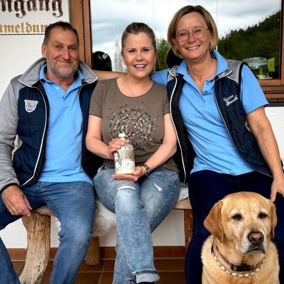 Drei Erwachsene und ein Hund sitzen lächelnd auf einer Bank draußen. Der Mann auf der linken Seite hält eine Flasche Vanillebier.