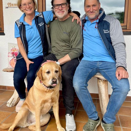 Drei Personen und ein Hund sitzen auf einer Bank, zwei Männer und eine Frau, der Hund sitzt vor ihnen.