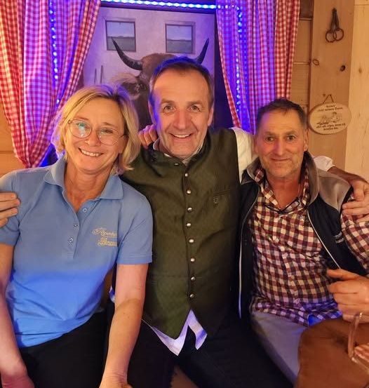 Drei Personen sitzen zusammen in einem Raum mit rustikalem Holzinterieur, eine Frau links, ein Mann in der Mitte mit seinem Arm um den Mann rechts.