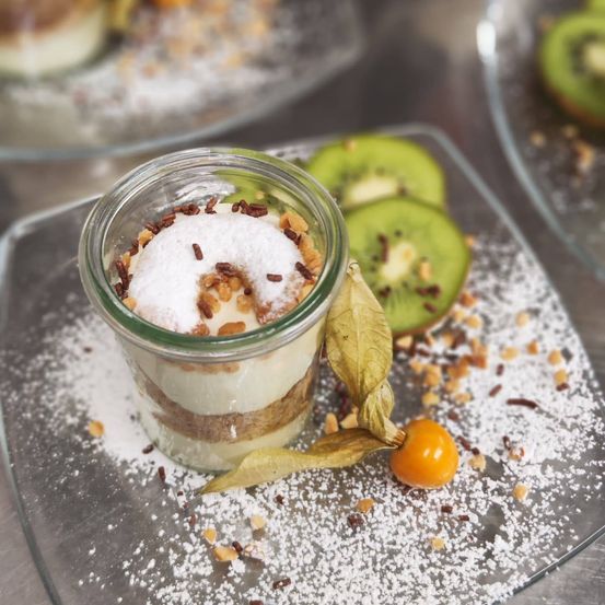 Ein Glasbecher mit geschichtetem Dessert, das mit weißer Pulver, Nüssen und Schokoladenstreuseln belegt ist. Es steht auf einem Teller mit verstreutem Pulver, Nüssen, einem Blatt und Kiwischeiben.