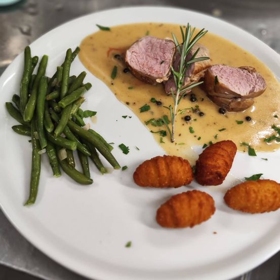 Ein Teller mit geschnittenen Schweinefilets in cremiger Sauce, garniert mit Rosmarin, grünen Bohnen und Kartoffelkroketten.