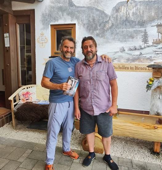 Zwei Männer stehen vor einem Gebäude. Einer hält ein Buch und beide lächeln. Hinter ihnen steht eine Bank, eine Wand mit einem Wandbild und eine Tür.