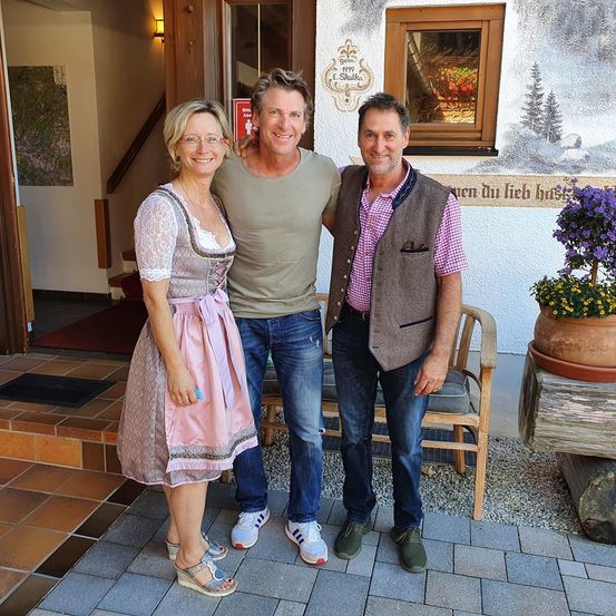 Drei Personen posieren für ein Foto vor einem Gebäude mit einer Wandmalerei, eine trägt ein rosa Kleid, eine andere ein graues T-Shirt und die dritte ein rosa und weißes Hemd.