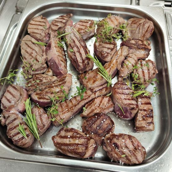 Ein Edelstahltablett gefüllt mit gegrilltem Fleisch, dekoriert mit Kräutern, wahrscheinlich Rosmarin und Thymian, präsentiert eine rustikale und appetitliche Anmutung.