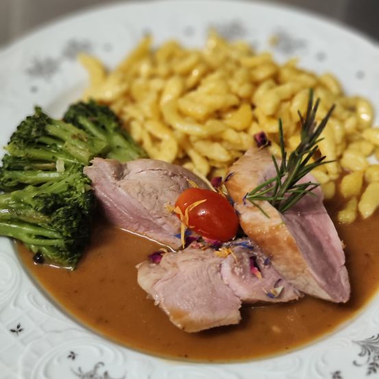 Ein Teller mit rosa Fleisch, Brokkoli und gelber Pasta, alles in brauner Sauce bedeckt. Ein Rosmarinzweig und eine Kirschtomate garnieren das Gericht.