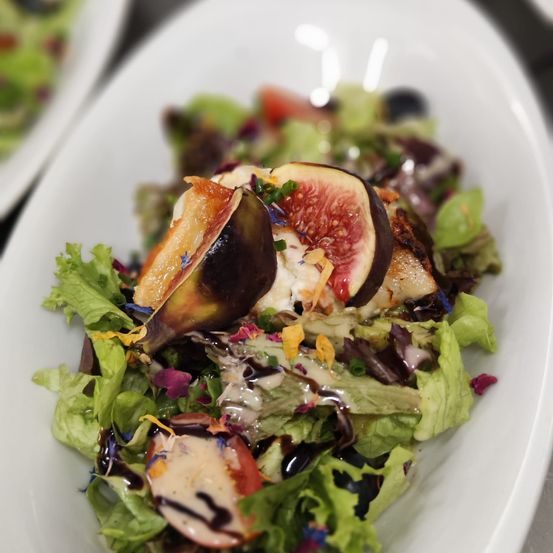 Ein weißer Teller mit einem farbenfrohen Salat, gekrönt von Feigen, Käse und einem Balsamico-Glasur. Der Salat besteht aus einer Mischung aus grünen und roten Blättern.