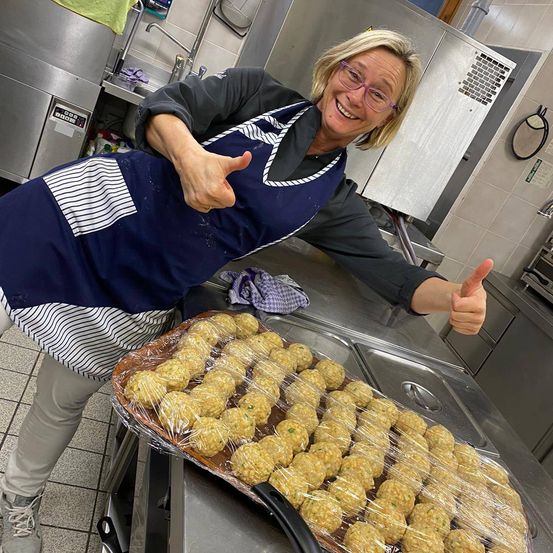 Eine Frau in einer Küche, die eine blaue Schürze trägt, lächelt und zeigt ein Daumen hoch, während sie eine Schale mit eingewickeltem Essen hält.