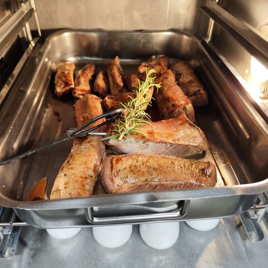 Ein silbernes Tablett hält mehrere Stücke gebratenes Fleisch, garniert mit Rosmarin. Ein Paar Zangen ruht auf dem Tablett. Das Tablett befindet sich in einem Ofen.
