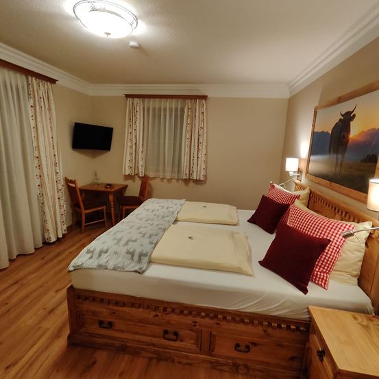 Ein gemütliches Schlafzimmer mit Holzboden verfügt über ein Bett, einen Schreibtisch und einen Fernseher. Die Wände sind mit Vorhängen und einem Bild von einer Kuh dekoriert.