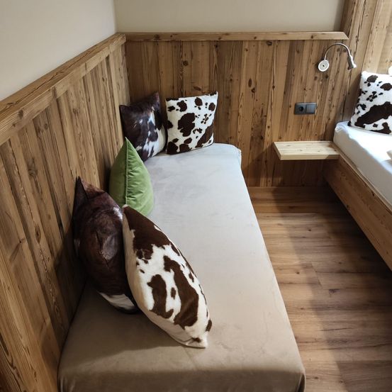 Eine gemütliche Ecke mit einem Einzelbett, bedeckt mit einem beigen Laken, und drei dekorativen Kissen. Holzwände und ein Holzboden vervollständigen das rustikale Interieur.