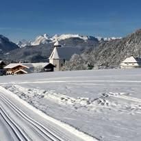 Eine verschneite Landschaft mit Bergen im Hintergrund, einer kleinen Kapelle und einem Haus, mit Spuren im Schnee.