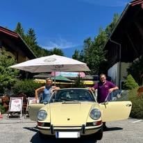 Zwei Männer stehen neben einem gelben Porsche 911 mit geöffneten Türen vor einem Haus mit einem weißen Sonnenschirm.
