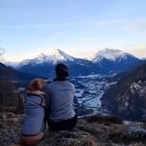 Eine Person und ein Hund sitzen auf einem Berggipfel und blicken auf die verschneiten Berge und ein Tal unten. Der Himmel ist blau mit einigen Wolken.