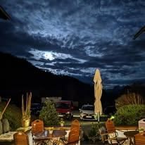 Die Außensitzfläche eines Restaurants unter einem Vollmond, mit geparkten Autos, Bergen in der Ferne und einem bewölkten Nachthimmel.