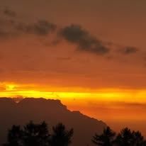 Sonnenuntergang über einer Bergkette mit einem warmen Schein, silhouettierten Bäumen und einem Himmel, der mit Orange- und Rosatönen bemalt ist.