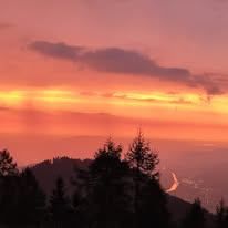 Ein Sonnenuntergangsblick von einem Berg mit leuchtenden lila und orangefarbenen Farbtönen, Bäumen und einem fernen Tal.