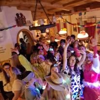 Eine lebhafte Gruppe von Menschen in einem festlichen Raum, einige in Kostümen, heben die Hände und benutzen Taschenlampen, mit Dekorationen, die von der Decke hängen.