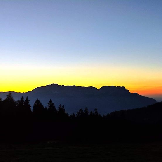 Sonnenuntergang über Bergen, Silhouette von Bäumen, leuchtendes orange und pink am Himmel, dunkler Vordergrund.