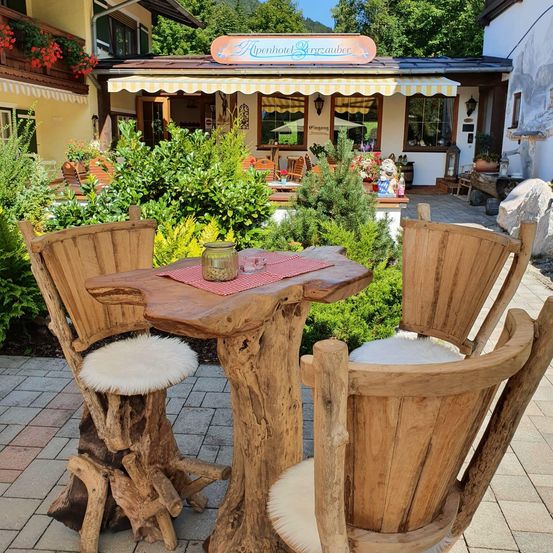 Ein Essbereich im Freien mit Holzstühlen und einem rustikalen Tisch vor einem gemütlichen Hotel mit blumengeschmücktem Balkon und gelbem Vordach.