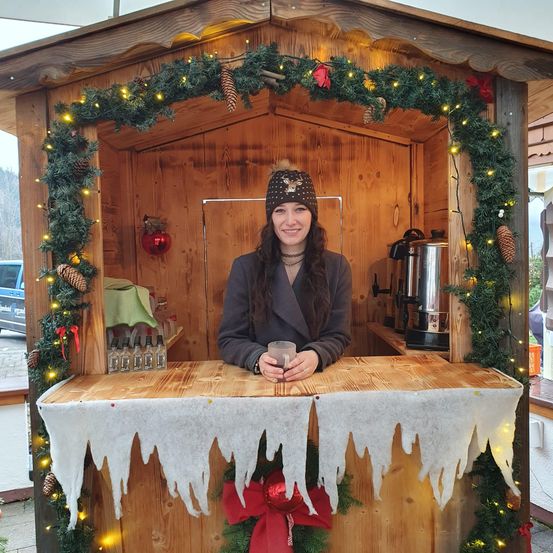 Eine lächelnde Frau steht hinter einem festlich geschmückten hölzernen Tresen und hält eine Tasse. Der Tresen ist mit Weihnachtslichtern und Grün geschmückt.