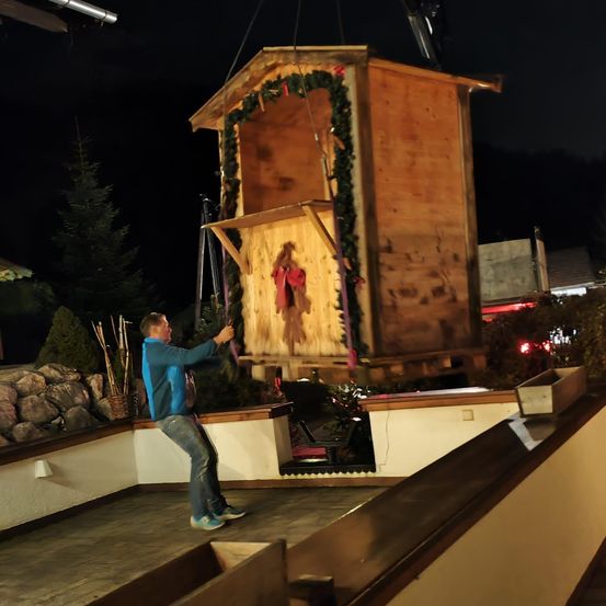 Ein Mann hebt nachts eine hölzerne Krippe. Die Szene ist mit Grünzeug dekoriert und hat ein rotes Band. Im Hintergrund befinden sich Bäume und Pflanzen.