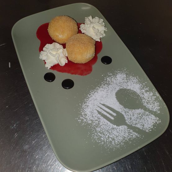 Ein Teller mit zwei frittierten Kugeln, überzogen mit Puderzucker, mit roter Sauce überzogen und mit Schlagsahne garniert. Eine Gabel und ein Löffel sind mit Puderzucker bestreut.