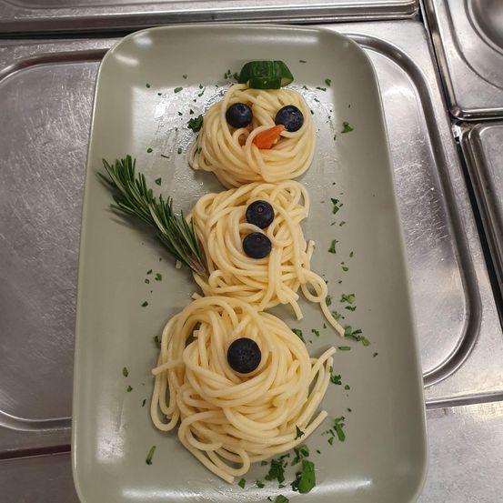 Ein Teller mit drei Nudelschneemännern. Die Schneemänner haben blaue Beeren als Augen, eine Karotte als Nase und Rosmarin als Arme. Der Teller steht auf einer Edelstahlfläche.