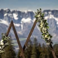 Ein mit weißen Blumen geschmückter Hochzeitsbogen steht im Vordergrund mit einer Gebirgslandschaft im Hintergrund.
