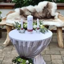 Ein weißer Tisch mit weißer Tischdecke, einer weißen Kerze und einem Blumenstrauß steht vor einer mit Fell bedeckten Holzbank.