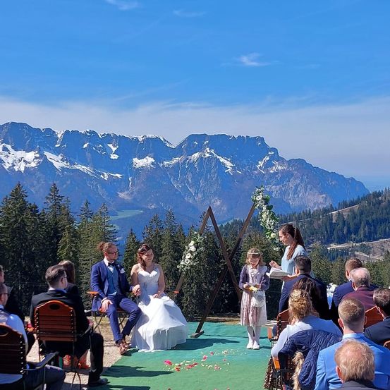 Eine Hochzeitszeremonie findet im Freien statt, mit einer Braut und einem Bräutigam auf Stühlen, umgeben von Gästen und einer wunderschönen Bergkulisse.