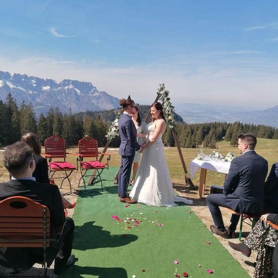 Eine Hochzeitszeremonie auf einem Grasfeld mit Bergen im Hintergrund. Die Braut und der Bräutigam stehen auf einem grünen Teppich mit Rosenblättern. Sie sind von Gästen umgeben, die auf Stühlen sitzen.