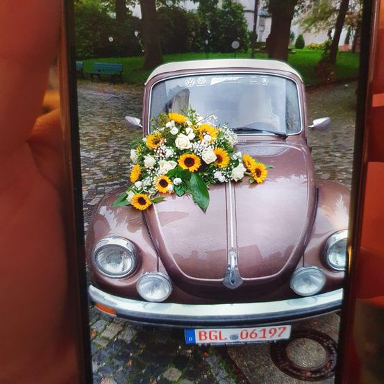 Ein brauner Oldtimer mit einem Strauß Sonnenblumen und weißen Blumen auf der Motorhaube, geparkt auf einer Kopfsteinpflasterstraße.