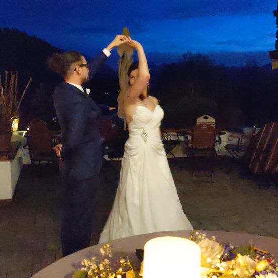 Ein frisch verheiratetes Paar tanzt bei Nacht auf einer Terrasse, mit einer Kerze und Blumen auf dem Tisch und einer Kulisse aus Bäumen und Bergen.