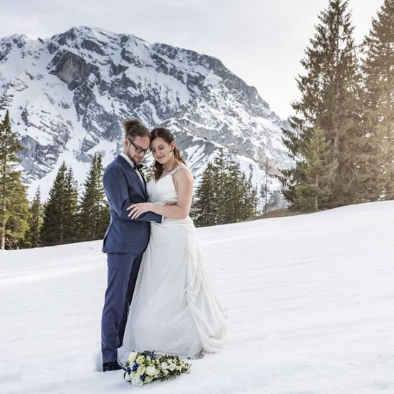 Ein frisch verheiratetes Paar steht auf einem verschneiten Berg, der Bräutigam in einem blauen Anzug und die Braut in einem weißen Hochzeitskleid. Sie sind von Kiefern umgeben und einer verschneiten Landschaft.