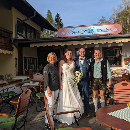 Eine Hochzeitsgesellschaft steht vor dem Alpenhotel Bergzauber, die Braut im Hochzeitskleid, der Bräutigam im blauen Anzug. Der Mann rechts trägt traditionelle bayerische Kleidung.