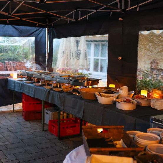 Ein Buffettisch ist unter einem Zelt aufgebaut, mit mehreren Gerichten und Schüsseln zur Schau gestellt. Der Tisch ist mit einem schwarzen Tuch bedeckt. Der Bereich ist schwach beleuchtet mit Kerzen auf dem Tisch.