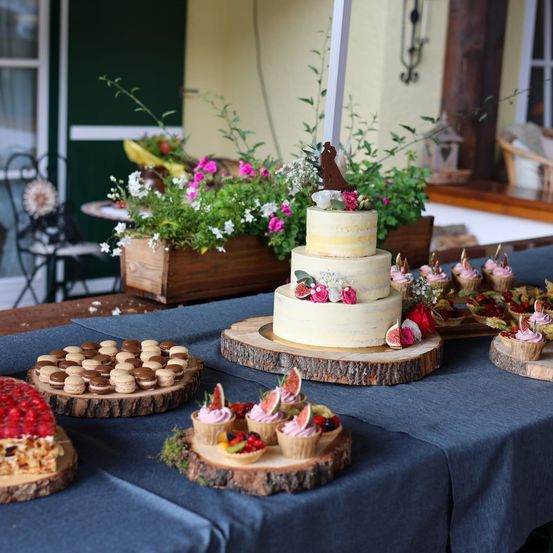 Ein Tisch zeigt einen großen dreistöckigen Hochzeitskuchen, Cupcakes und Macarons. In der Nähe steht ein hölzerner Blumenkasten mit rosa und weißen Blumen. Die Umgebung umfasst einen Stuhl und ein Haus im Hintergrund.