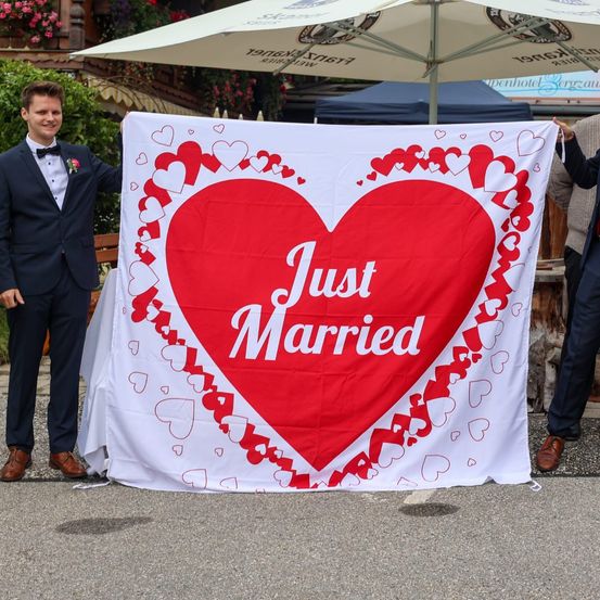Ein Mann in einem Anzug hält ein herzförmiges Banner mit der Aufschrift 'Just Married'. Er steht unter einem Regenschirm.