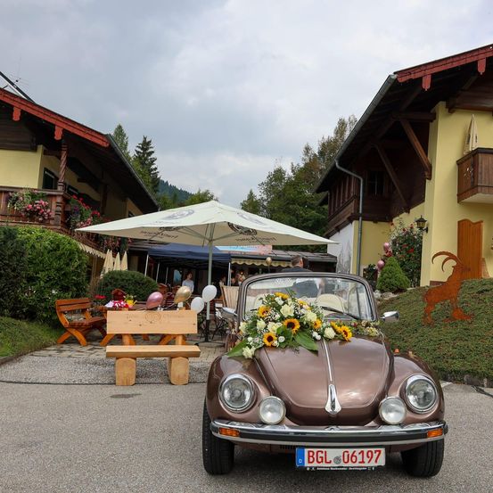 Ein alter VW Käfer mit Blumen ist vor einem Chalet geparkt, unter einem Regenschirm. Eine Bank und ein Tisch sind in der Nähe. Eine Hirschstatue steht am Hang.