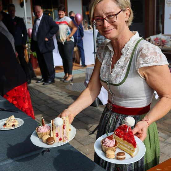 Eine Frau mit Brille und einem weißen Oberteil hält zwei Teller mit Desserts, darunter ein Erdbeerkuchen und Macarons, bei einer Veranstaltung im Freien.